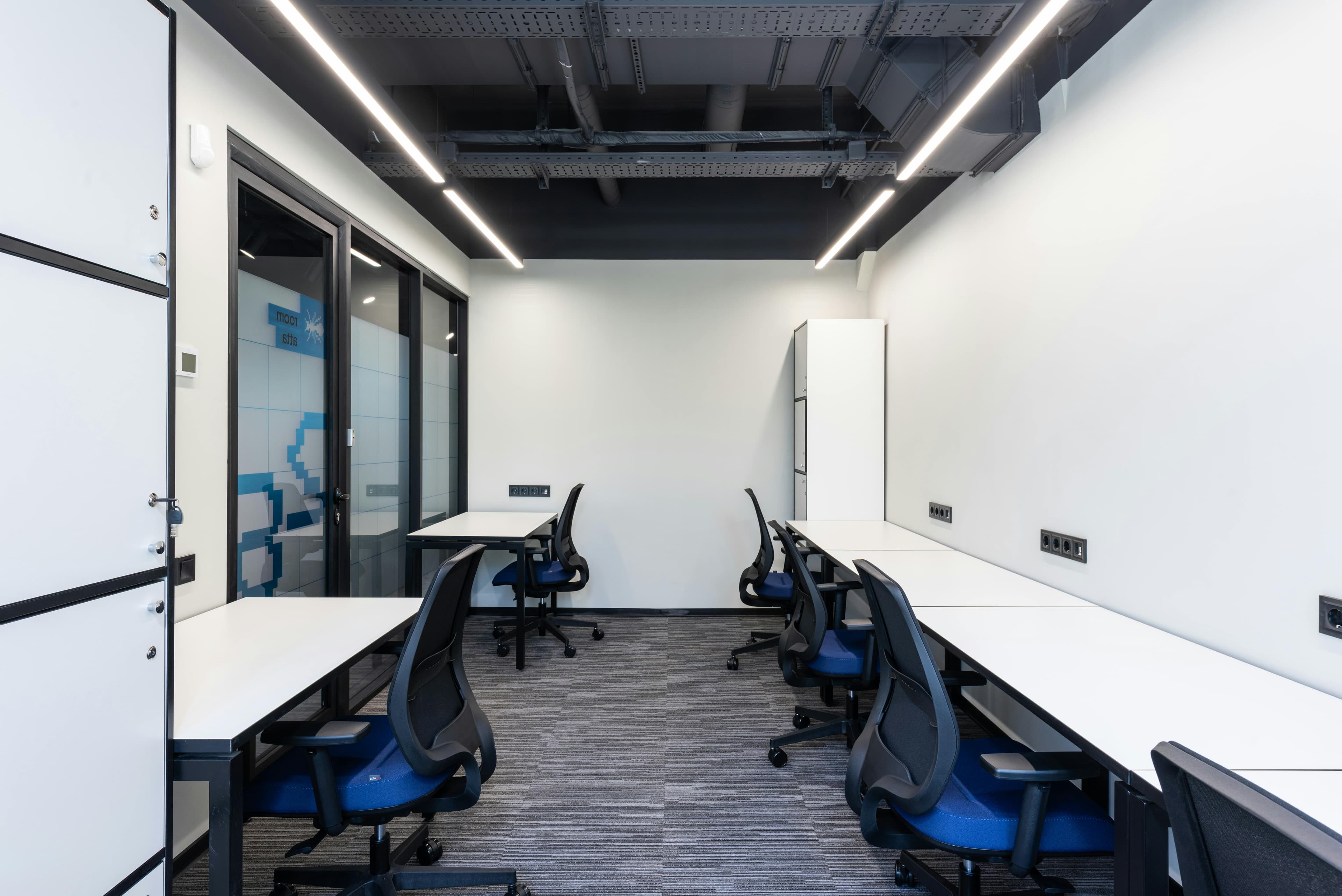 Modern office with white desks, blue ergonomic chairs, and industrial black ceiling with LED lights.