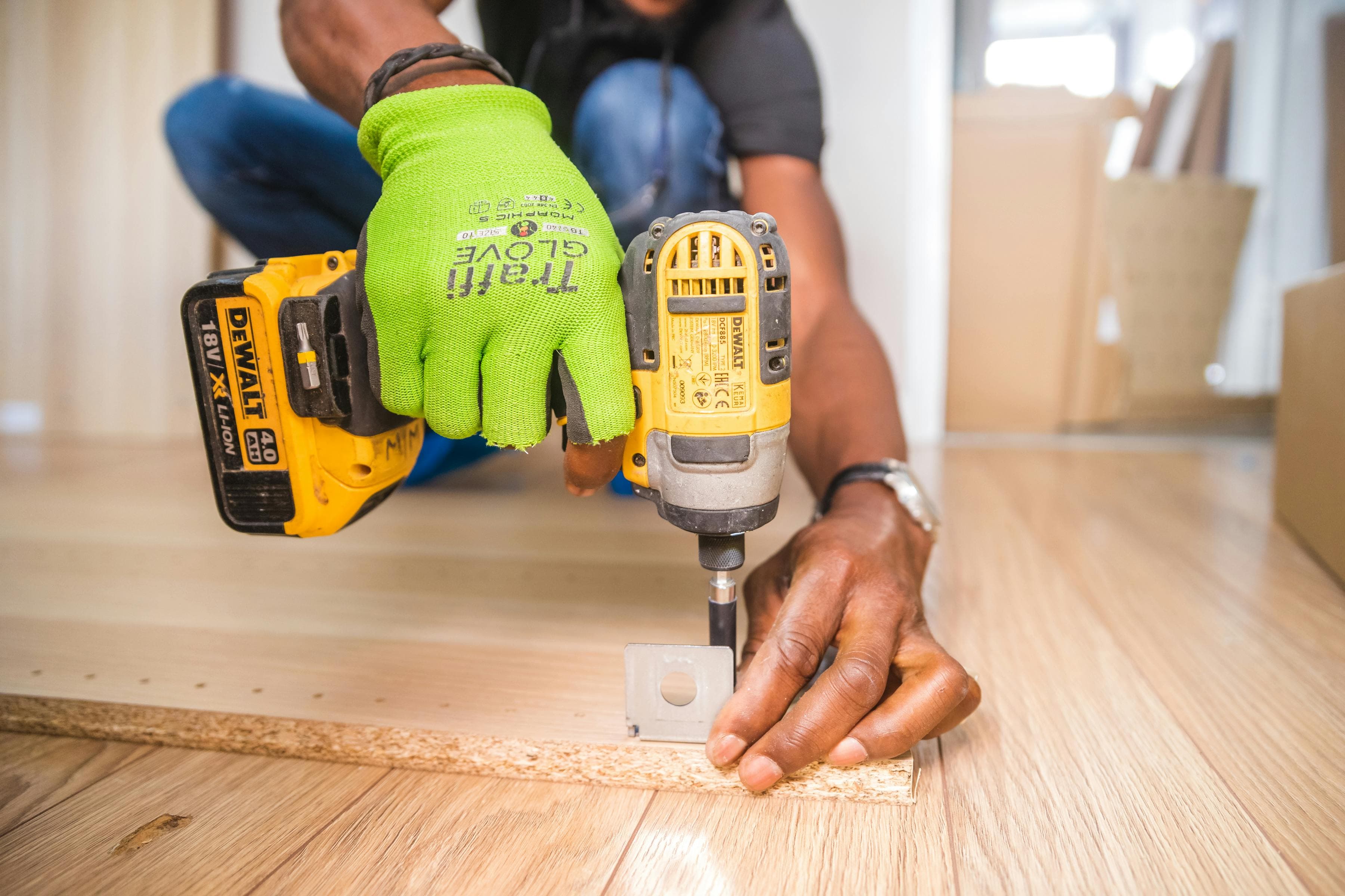 Hands using a yellow DeWalt drill to assemble furniture on a light-colored wooden floor.