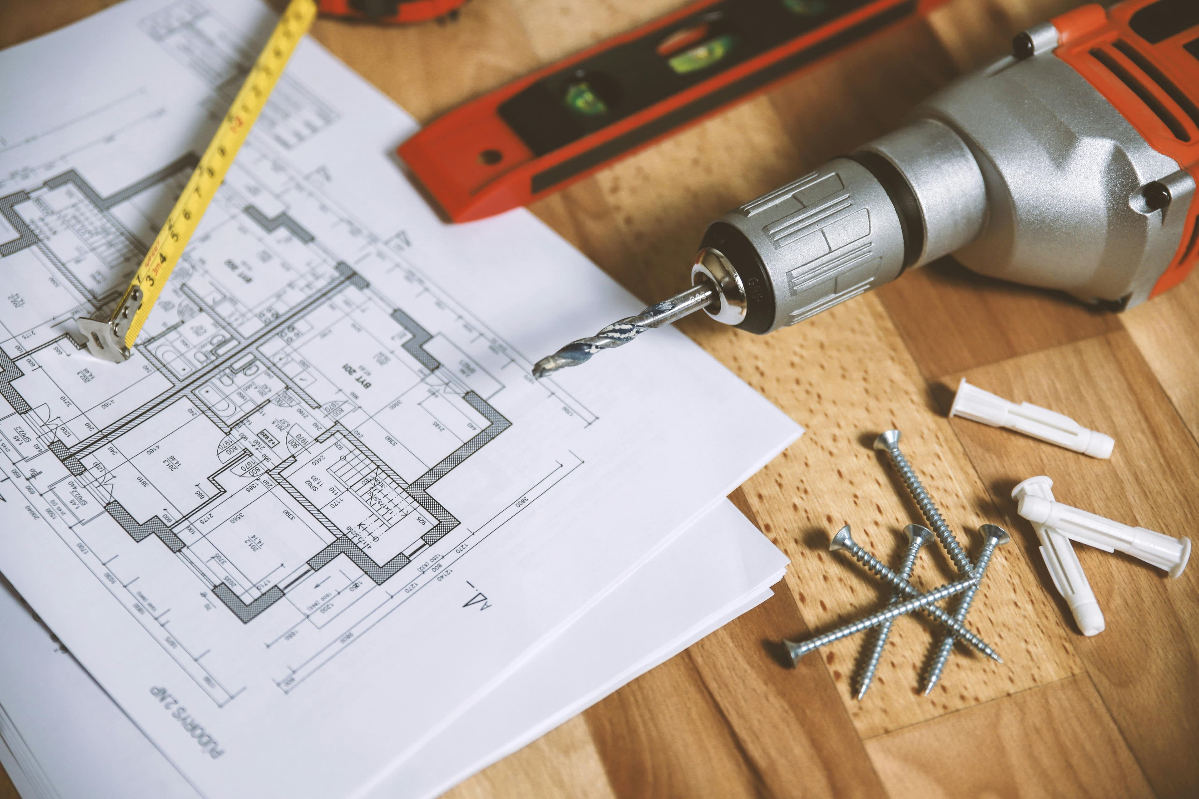 Electric drill, measuring tape, and screws scattered over architectural blueprints on a wooden surface.