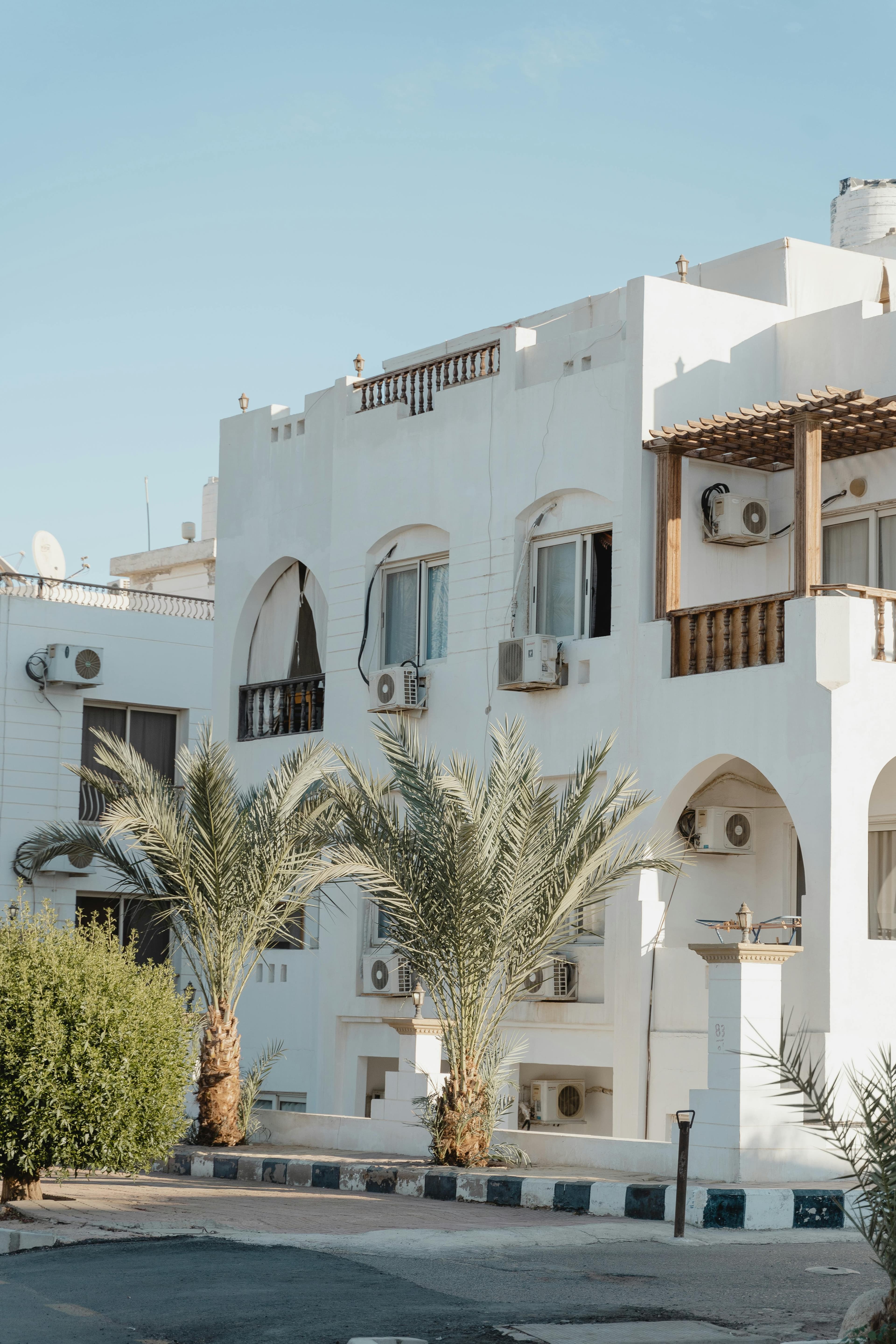 White Mediterranean-style building with arched windows and air conditioners behind two green palm trees.