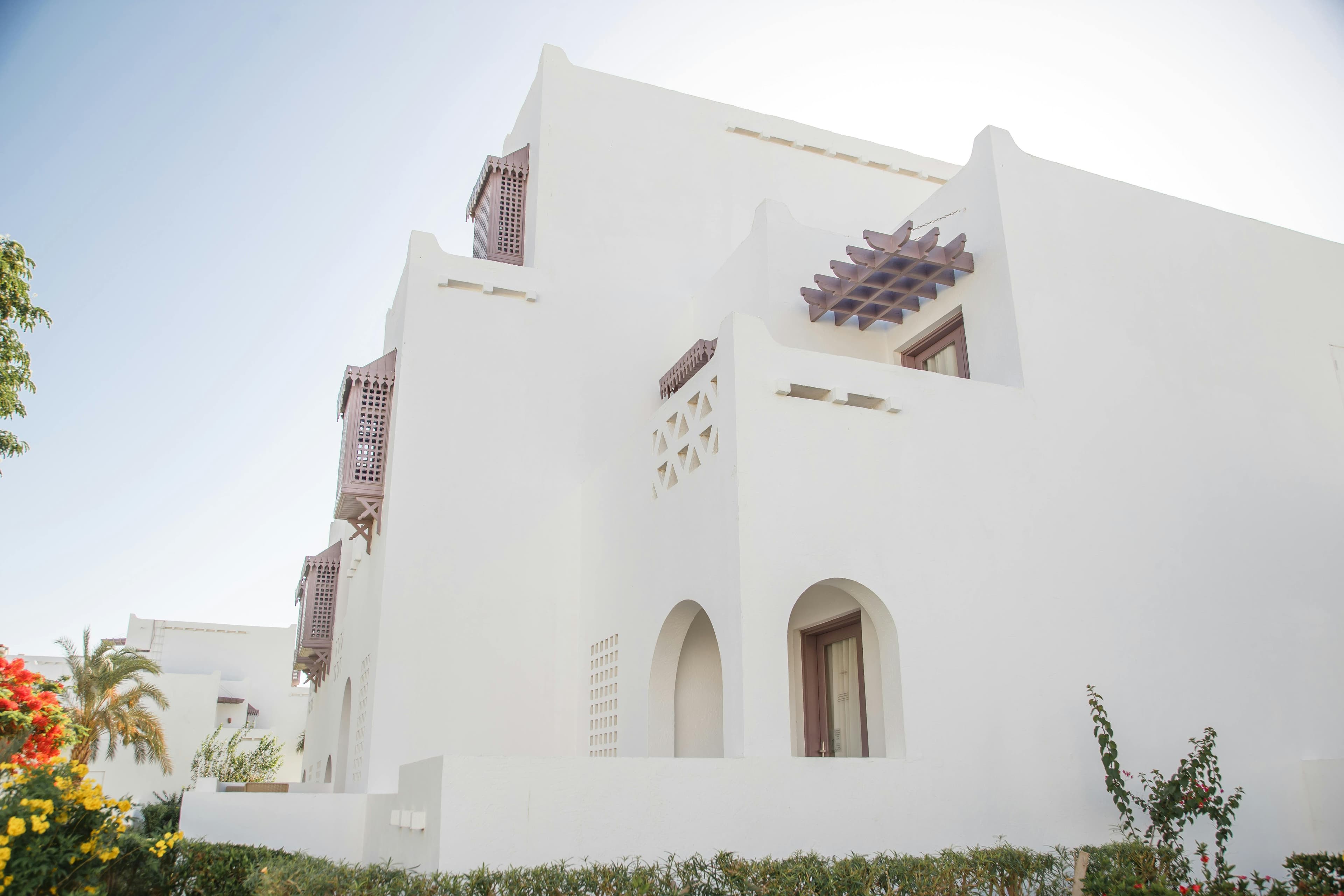 White building with traditional wooden lattice windows, arched doorways, and lush green garden landscaping.